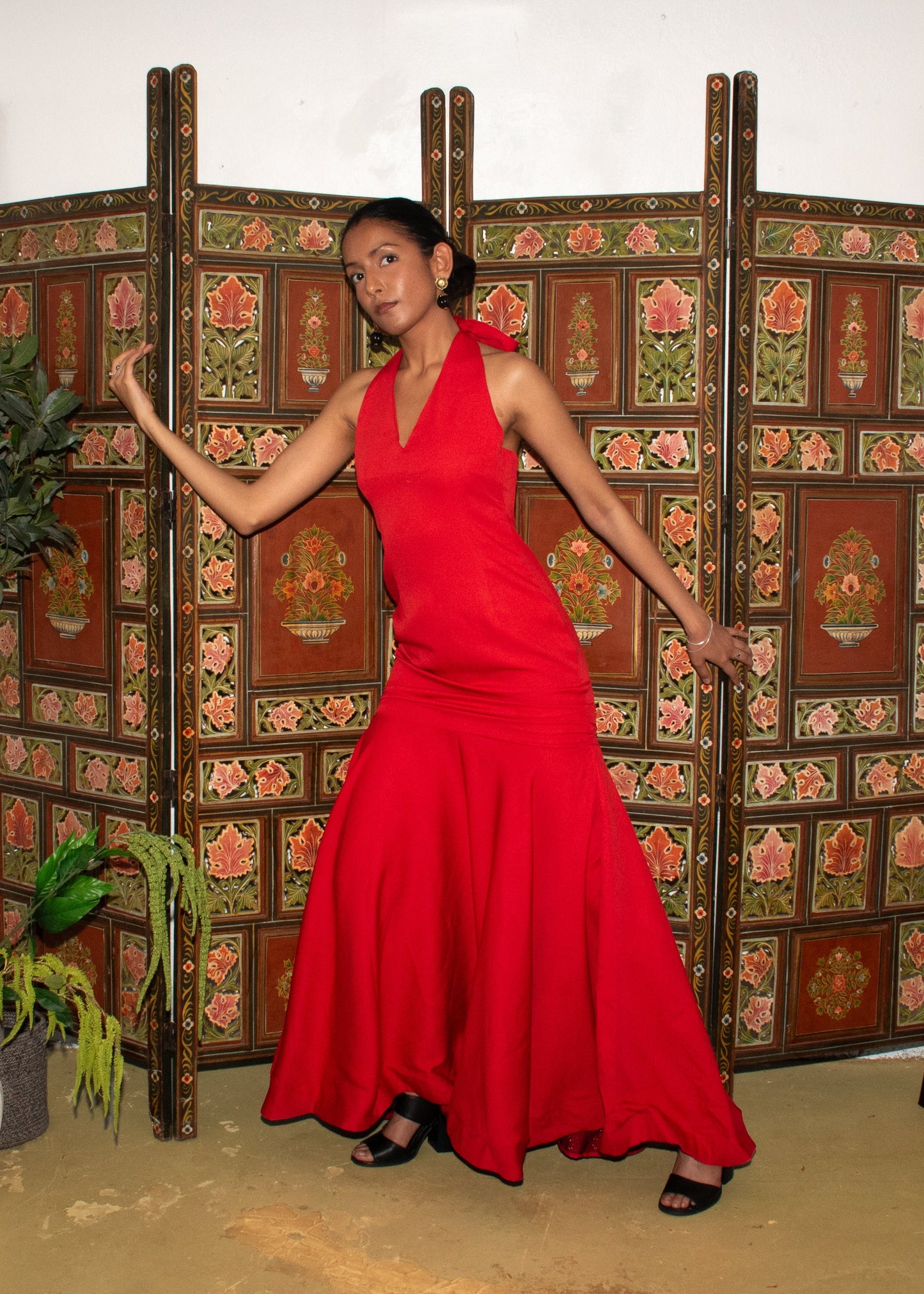 A woman wearing a sleeveless, vibrant red maxi dress stands in front of a decorative folding screen in an indoor room with white walls, beige carpet, a potted plant, and framed artwork.